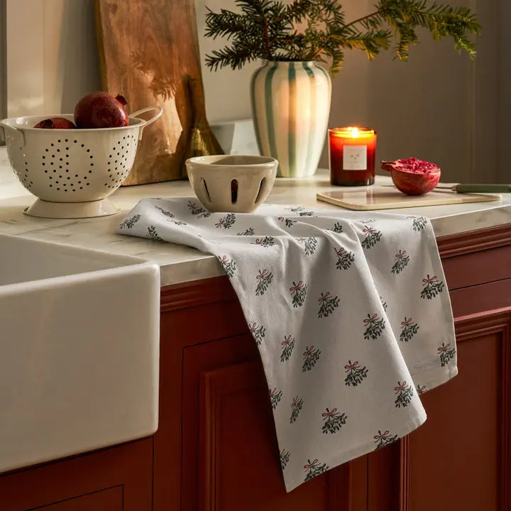 White tea towel with repeated mistletoe pattern draped over a bench with bowls and a candle
