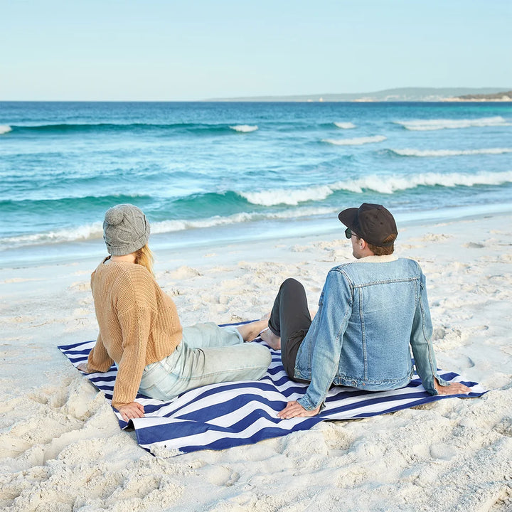 Two people sitting on a blue and white striped beach blanket on a sandy beach facing the ocean.