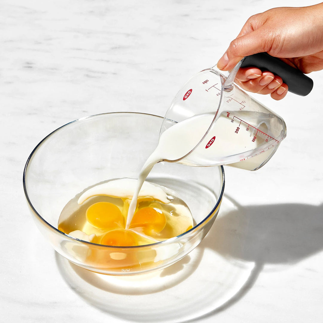 Person pouring milk from a measuring cup into a bowl with eggs on a light background