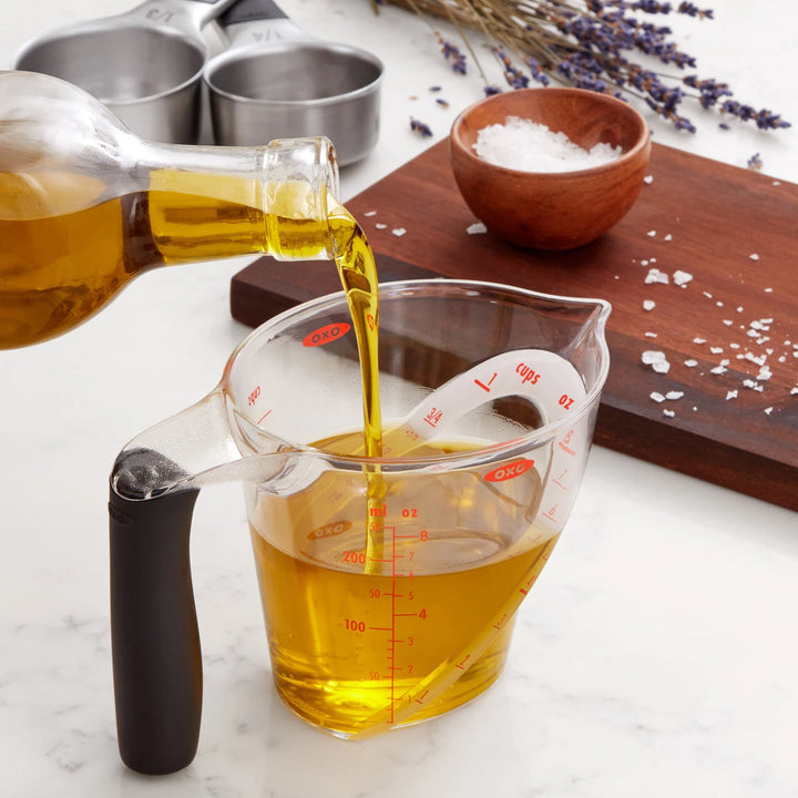 Oil being poured from a bottle into a measuring cup on a marble surface with lavender flowers.