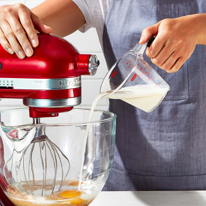 Person using a red KitchenAid mixer with a clear bowl, pouring liquid into it.