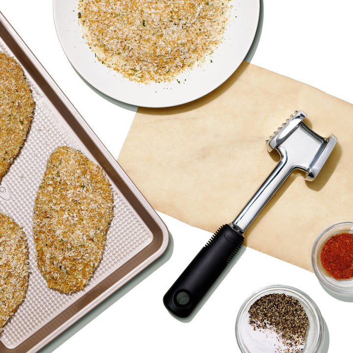 Breaded food items on a baking tray with a tenderizer and spices on a white background