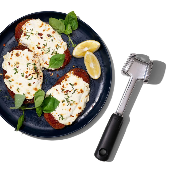 Plated dish of meat with cheese and herbs, accompanied by a meat tenderizer on a white background.