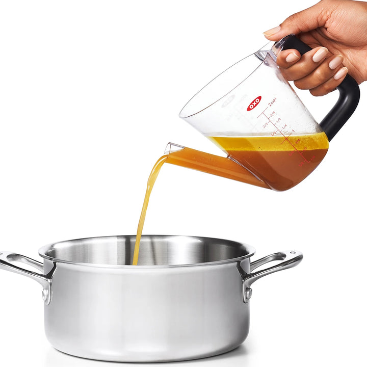Hand pouring oil from a measuring cup into a pot on a white background