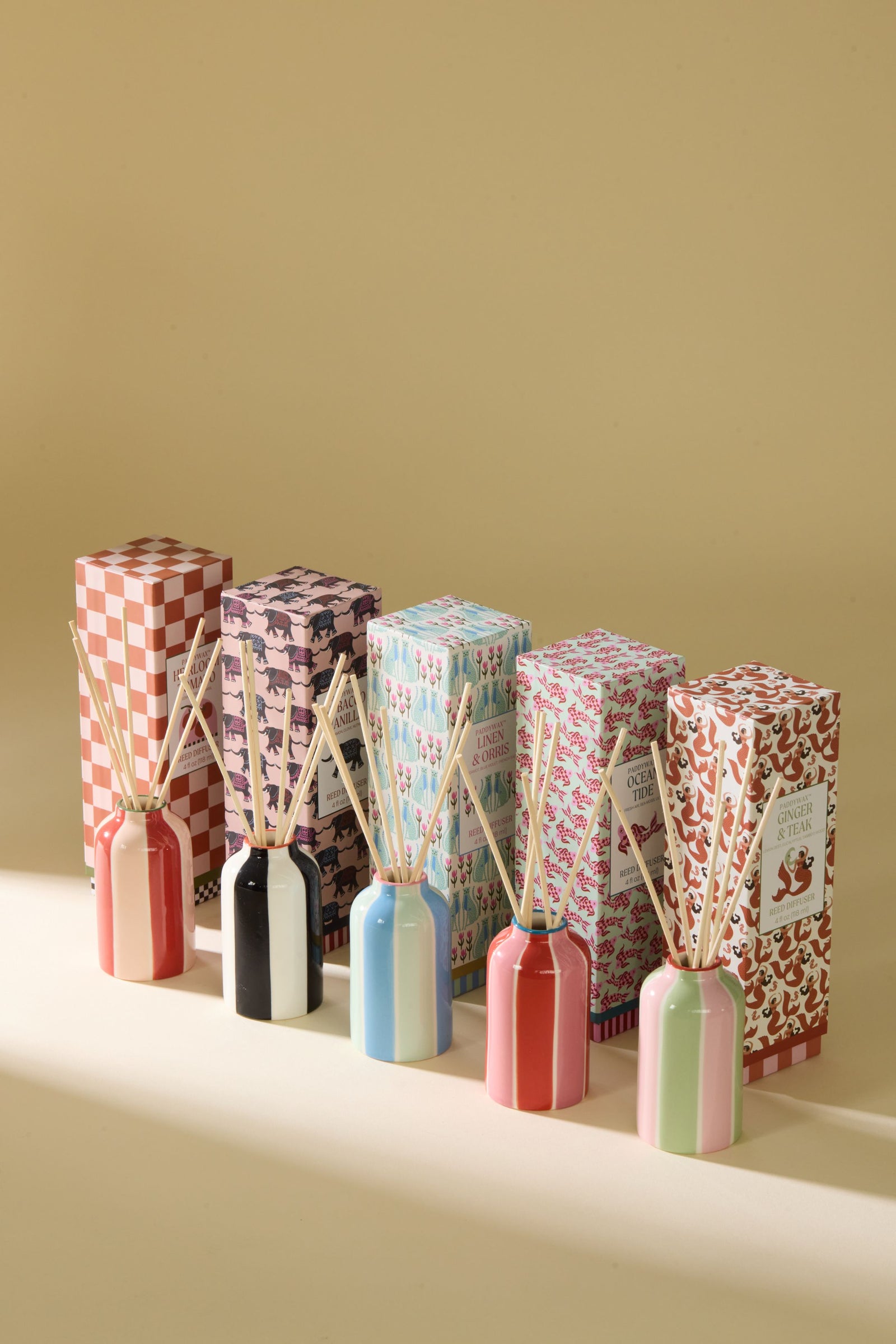 Six reed diffusers with colorful patterned boxes and wooden reeds, arranged in a row on a beige surface against a warm beige background.