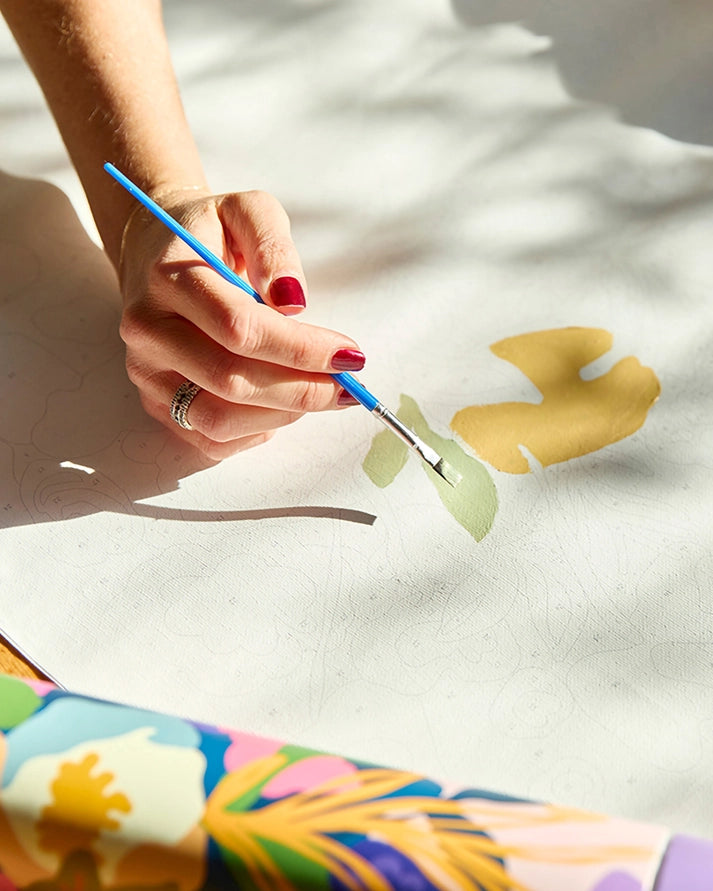 A close-up of a hand with red nail polish holding a blue-handled paintbrush and painting on a white canvas. A colorful tropical print fabric is partially visible in the lower foreground.