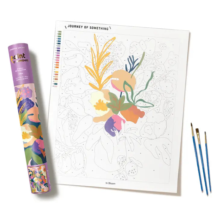 A floral paint by numbers named In Bloom, featuring a printed painting page with a partially colored flower bouquet, a purple branded coloring tube, and two blue paintbrushes on a white background.