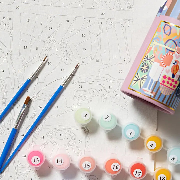 A flat-lay view of a DIY canvas paint-by-numbers kit, featuring a pink cup filled with colorful paint, multiple numbered paint pots, two blue paintbrushes, and a printed canvas sheet with numbered sections.