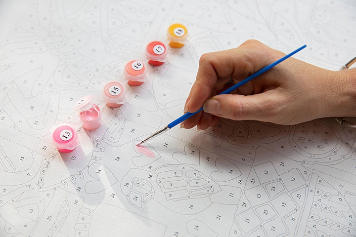 Hand holding a paintbrush with small containers of paint on a white surface with patterns.