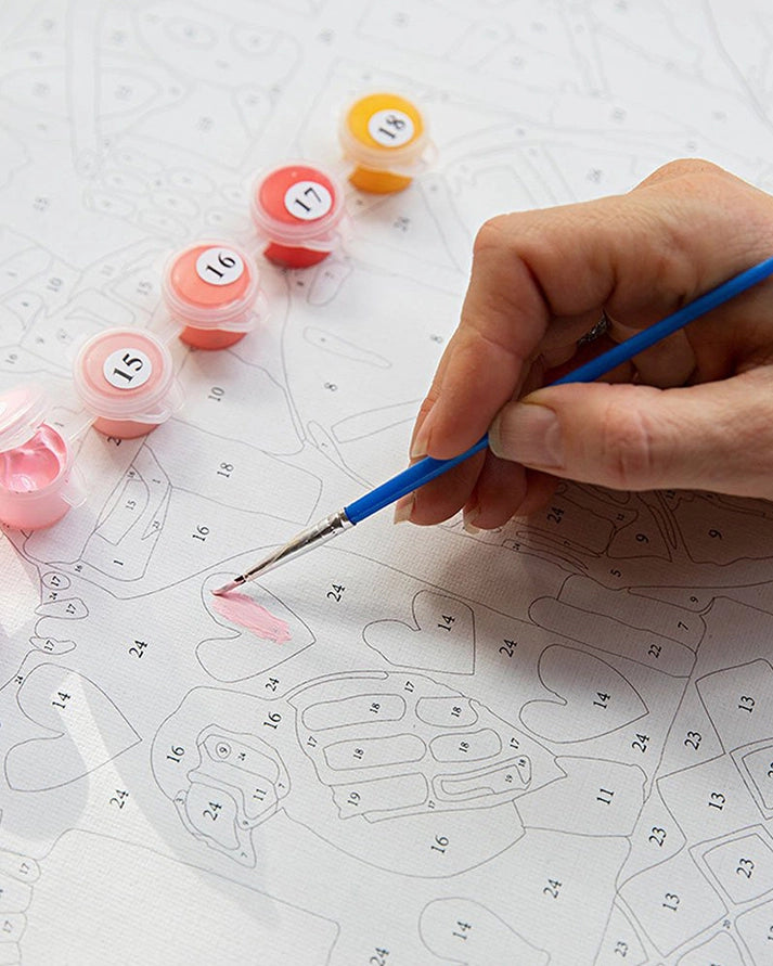 A hand holding a blue paintbrush applies pink paint to a numbered canvas while several small paint pots in pink, coral, orange, and yellow sit nearby on a white surface.