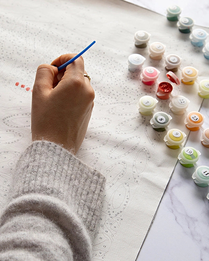 Hand holding a paintbrush near small containers of paint on a textured surface.