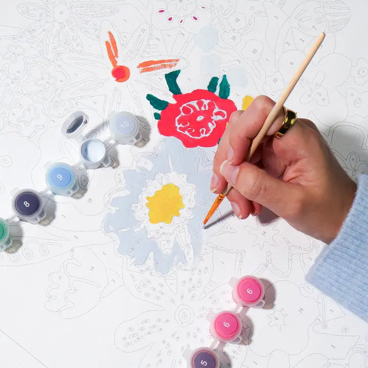 A hand holding a paintbrush painting a floral design on a white canvas, surrounded by small paint pots in various colors.