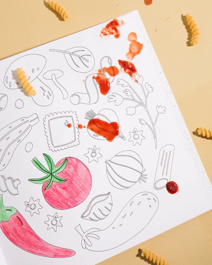 A flat-lay of a white coloring sheet featuring black outline drawings of vegetables and food items, with some sections colored in with red and pink paint and pasta pieces scattered around on a beige background.