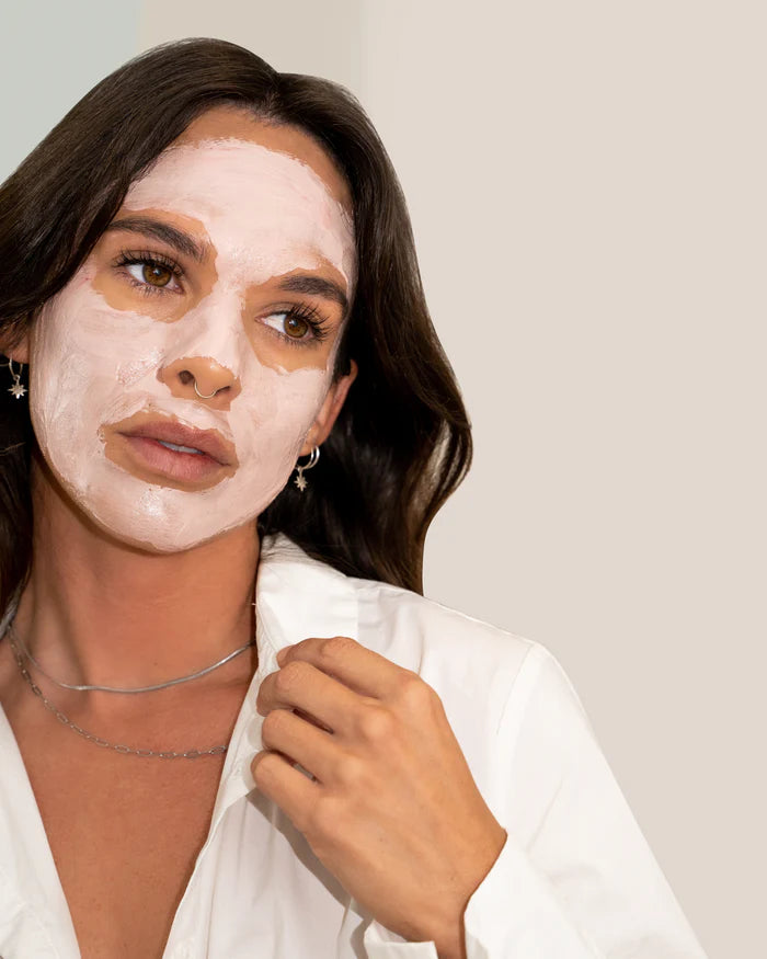 A woman with  a white shirt on and a clay facial mask on her face