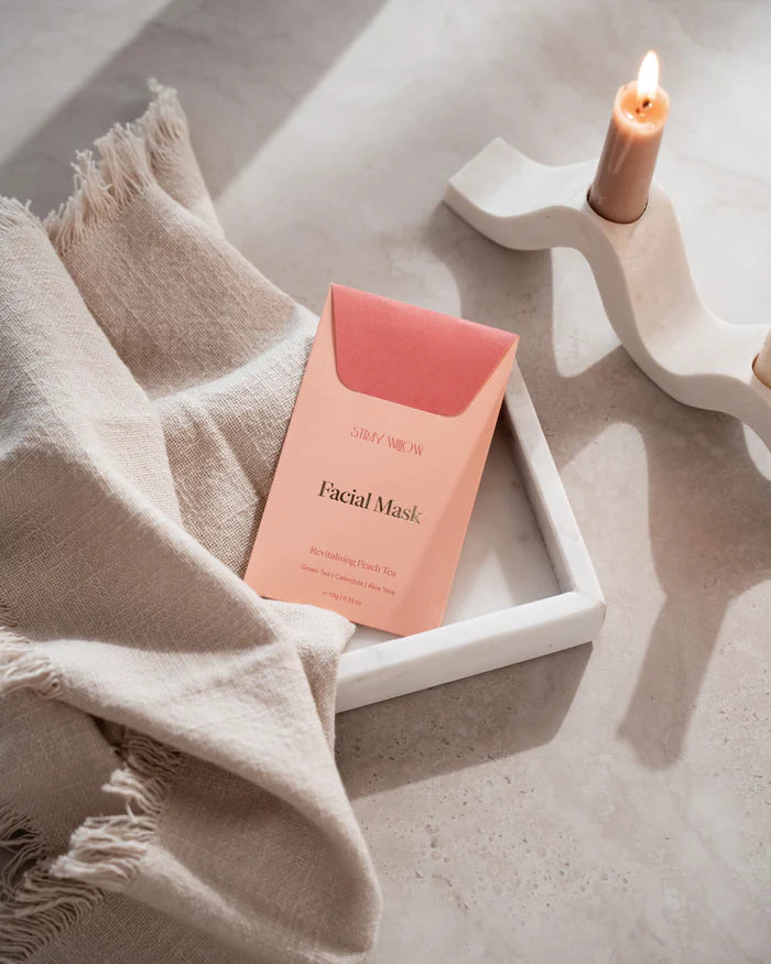 Facial mask packaging on a white tray with a candle and beige towel in the background