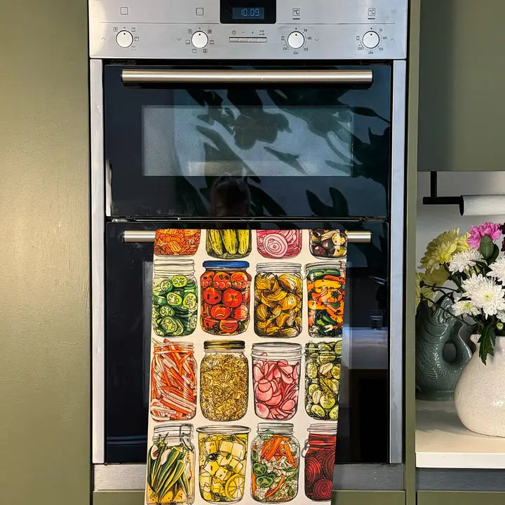 A stainless steel built-in oven with a glass door and a colorful kitchen towel hanging from the handle. The towel features illustrations of jars filled with sliced vegetables like cucumbers, tomatoes, onions, and carrots.