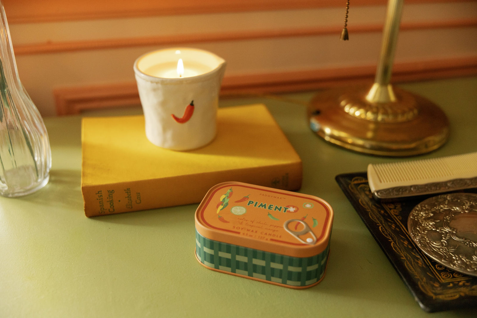 A styled tabletop scene with a small rectangular tin candle labeled "PIMENT" in the foreground, a lit white candle in a ceramic holder on a yellow book, and other decor objects in a warm indoor setting.