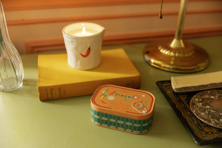 A styled tabletop scene with a small rectangular tin candle labeled "PIMENT" in the foreground, a lit white candle in a ceramic holder on a yellow book, and other decor objects in a warm indoor setting.