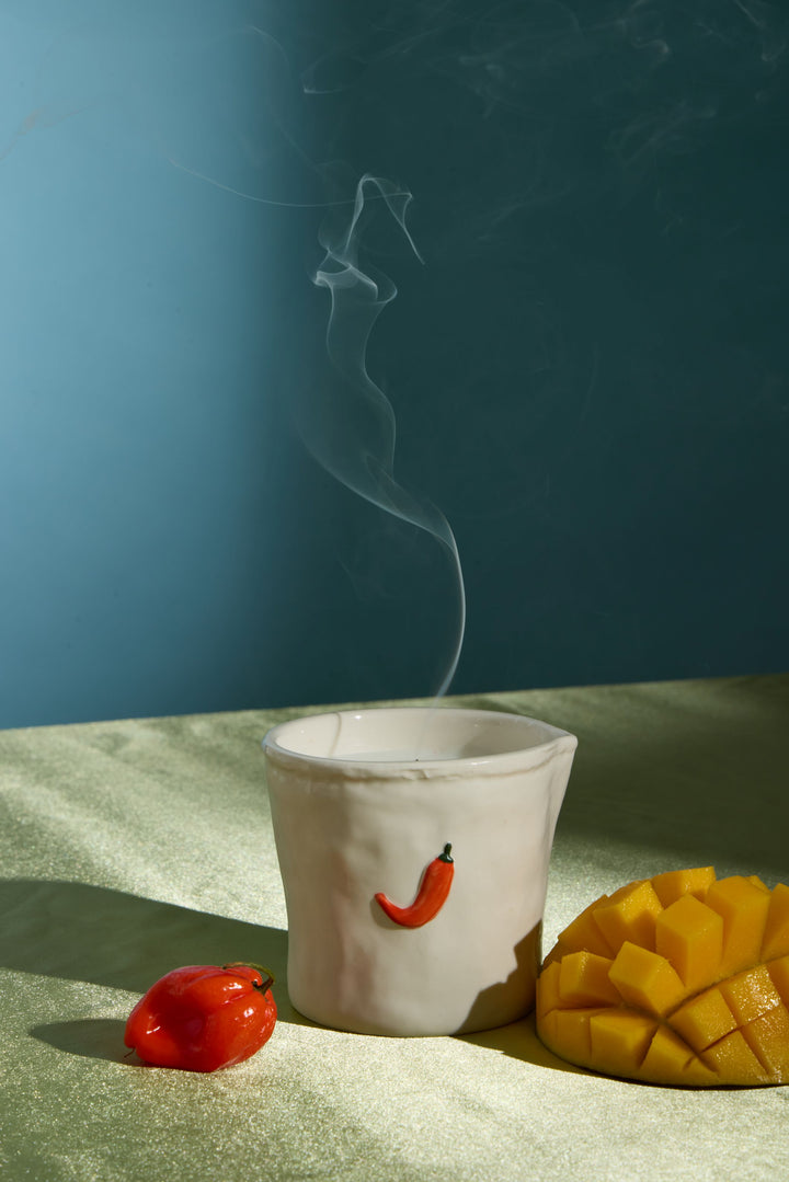A white ceramic mug with a small red chili pepper decoration, shown with steam rising from it on a textured beige surface. A real red chili pepper and a sliced mango are placed beside the mug against a blue background.