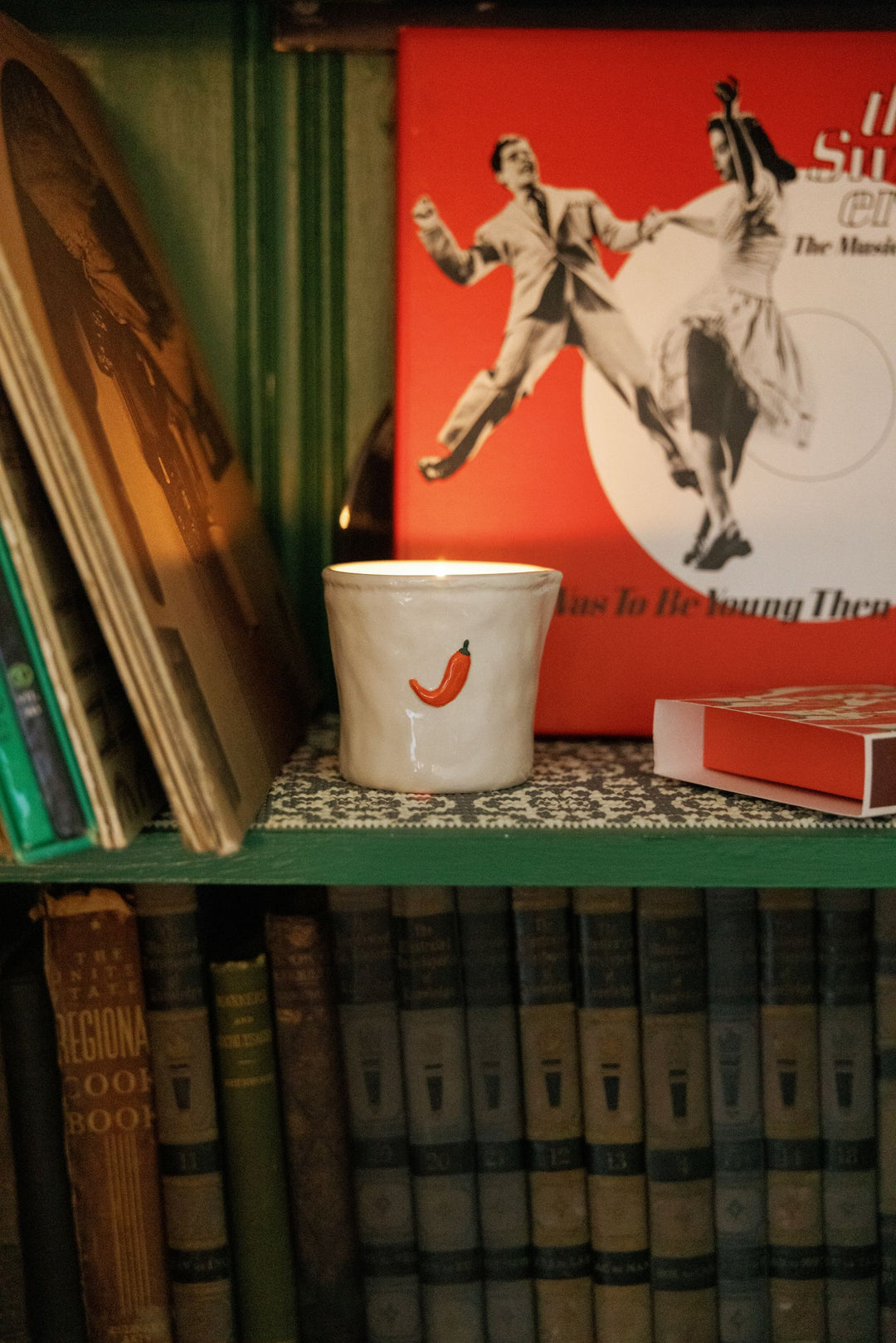 A white ceramic mug with a small red chili pepper graphic sits on a green bookshelf surrounded by vintage books and a red poster.