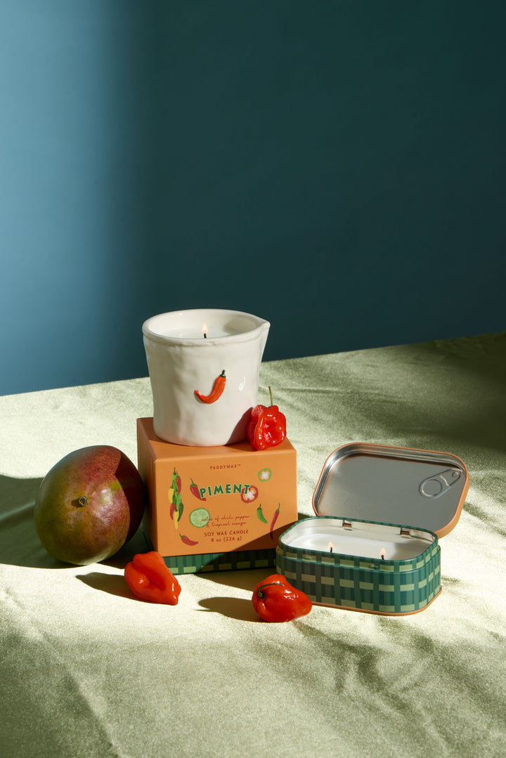 A styled product shot of a candle set on a draped fabric surface against a dark blue wall. The set includes a white ceramic candle vessel with a red chili pepper illustration, an orange box labeled &