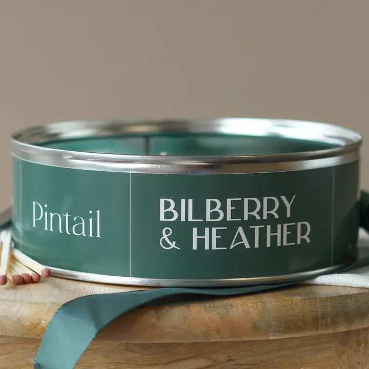 Green candle tin with 'Pintail Bilberry & Heather' text on a wooden surface.
