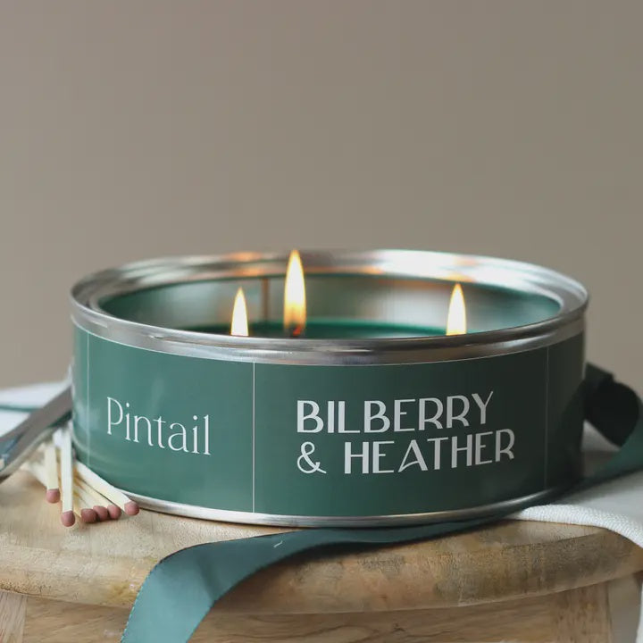Candle in a green can labeled 'Bilberry & Heather' by 'Pintail' on a wooden surface.
