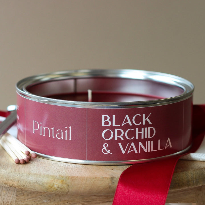 A maroon paint tin candle with Pintail Black Orchid &amp; Vanilla text on it on a wooden stool with matches and a red ribbon
