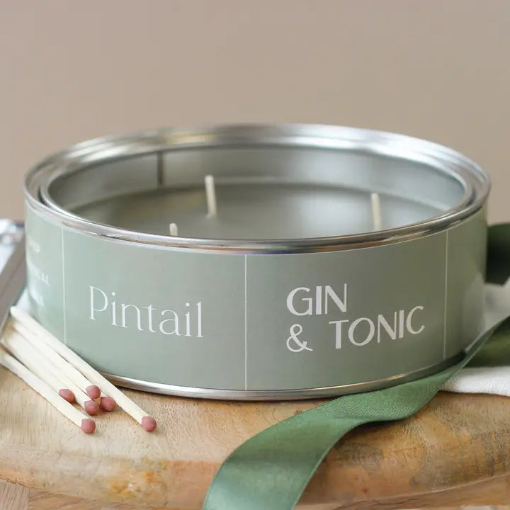 Candle in a metal tin labeled 'Pintail Gin & Tonic' with matches and a green ribbon on a wooden surface.