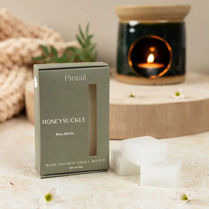 Box of Pintail Honeysuckle wax melts on a surface with a lit candle in the background.