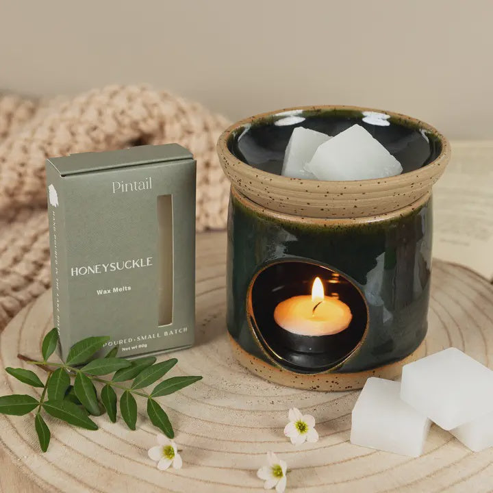 Ceramic wax melt burner with a lit candle, surrounded by honey-suckle wax melts packaging and decorative elements on a wooden surface.