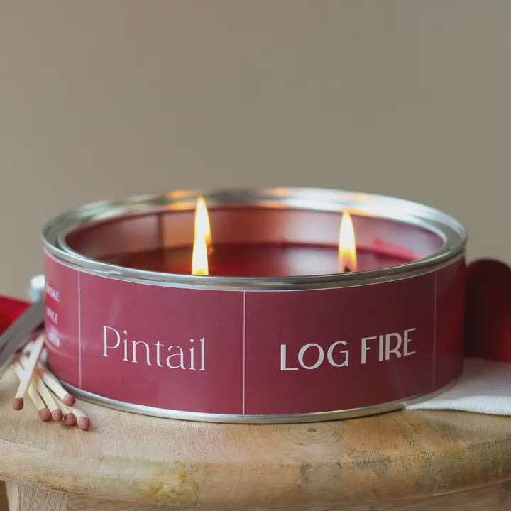 Candle in a red can labeled 'Pintail Log Fire' with matches on a wooden surface.