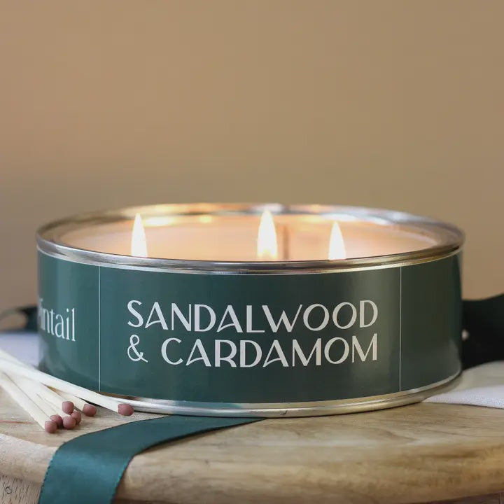 Sandalwood &amp; Cardamom candle in a green tin with wooden wick trimmer on a wooden surface.