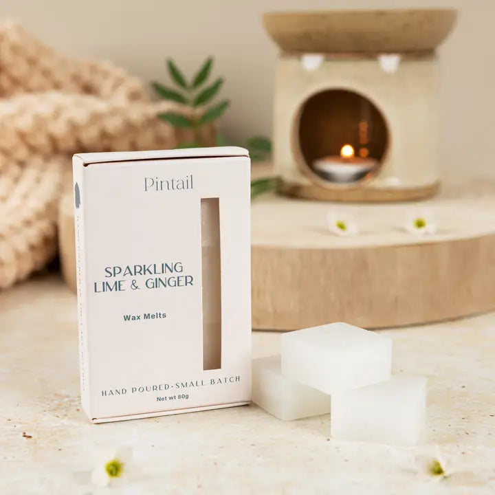 Pintail sparkling lime &amp; ginger wax melts packaging on a wooden surface with a candle in the background.