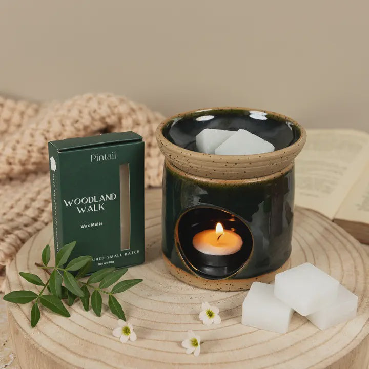 Ceramic wax melter with a lit candle, surrounded by wax melts and a 'Woodland Walk' packaging on a wooden surface.