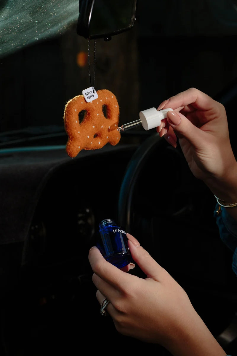 A hand putting fragrance on a car air freshener in the shape of a pretzel
