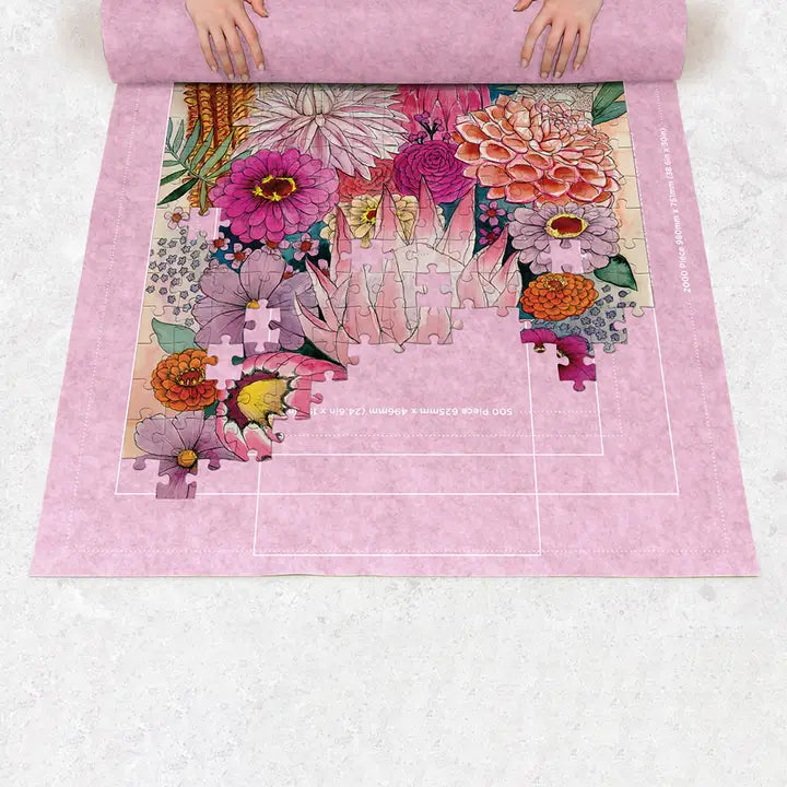 A pink jigsaw puzzle being unrolled, showing a floral illustration with partial puzzle pieces placed over the center. Hands are visible at the top holding the rolled edge, and the background is a light stone surface.