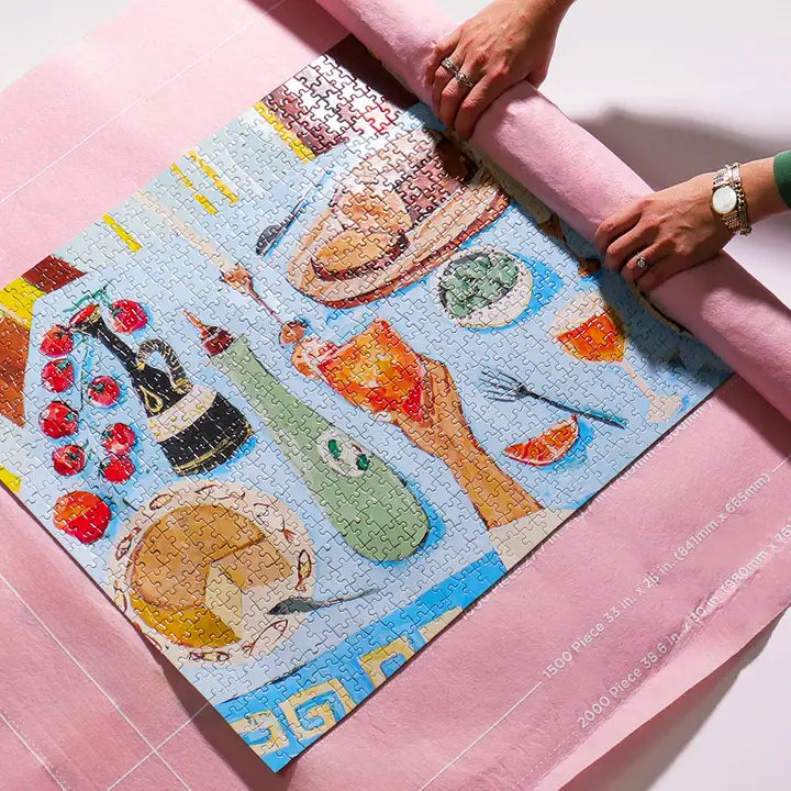 A jigsaw puzzle featuring a colorful Mediterranean-themed still life painting, being unrolled by two hands over a pink puzzle mat.