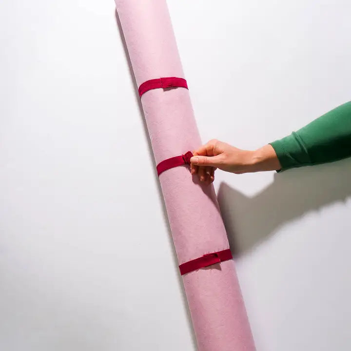 A hand in a green sleeve holds a pink rolled jigsaw puzzle mat against a plain white wall, secured with three red bands.