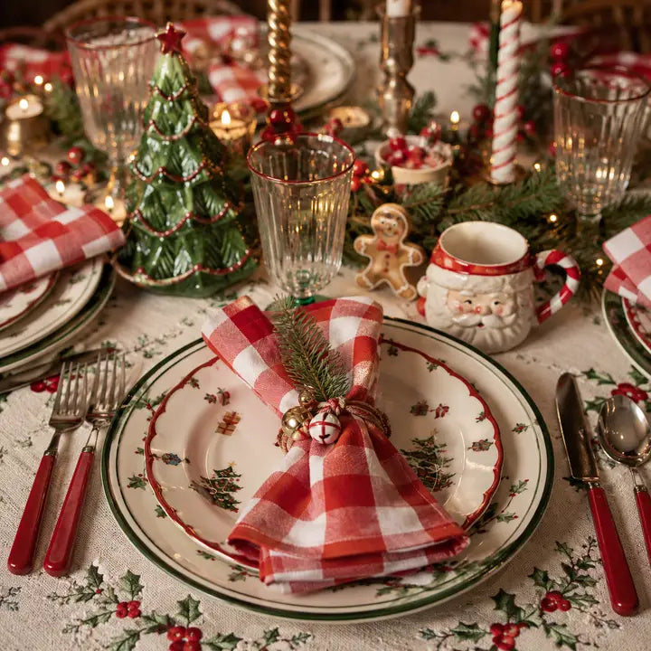 A festive Christmas table setting featuring a ceramic plate with a green and red holly border, a red and white checkered napkin tied with a pine sprig and gold bead, matching red-handled cutlery, and holiday decorations including a small green tree, candlesticks, and a Santa mug.