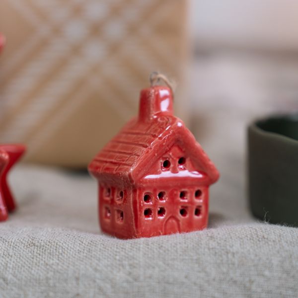 A small glossy red ceramic house ornament with a hanging loop, photographed on a textured fabric surface with a softly blurred background.