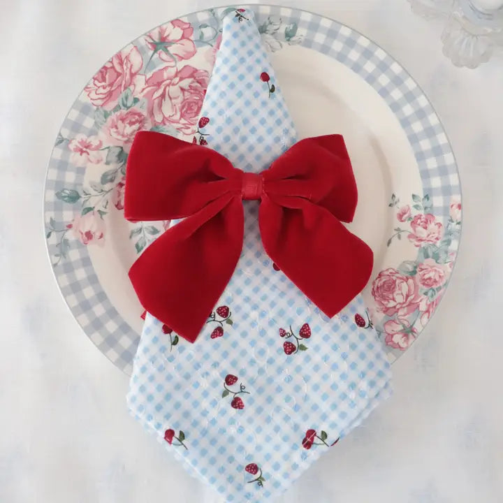 A folded light blue gingham napkin with a small red rose and berry print, tied with a large red velvet bow, placed on a white plate with a blue gingham border and pink floral print.