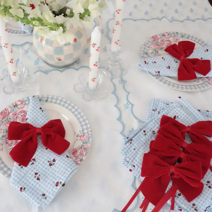 A styled tabletop setting with a white tablecloth, floral plates, white candles, and folded napkins tied with large red velvet bows. The napkins are light blue and white gingham with small red floral details.