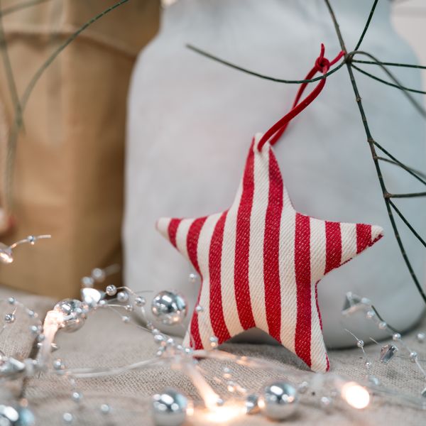 A fabric star-shaped Christmas ornament with red and white vertical stripes, hanging from a red ribbon against a softly blurred holiday background with lights and silver ornaments.