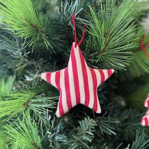 A fabric star-shaped Christmas ornament with red and white vertical stripes hanging from a red string on a green artificial Christmas tree.