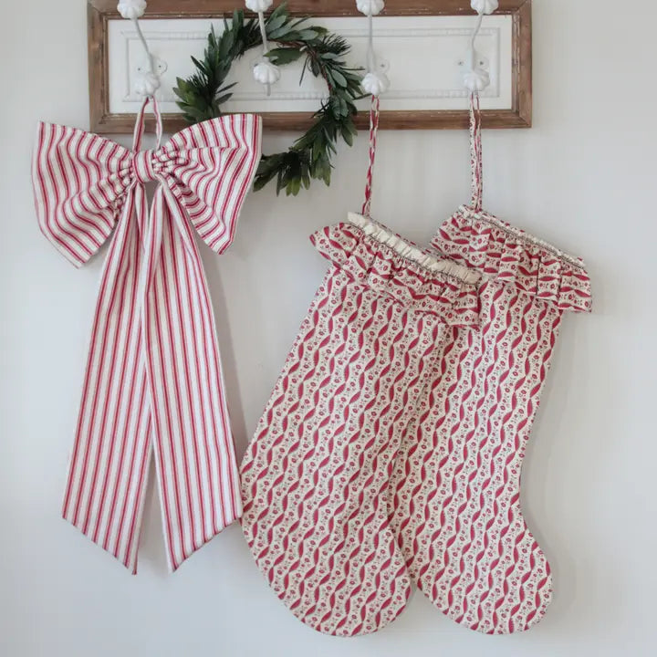 A Christmas-themed fabric hanging set featuring a large red and white striped bow with long tails and two matching red and white patterned stockings, displayed on a white wall hook rack with a green wreath.