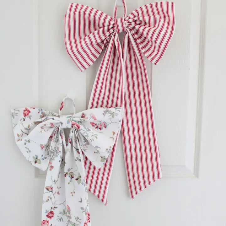 Two fabric bows hanging on a white door. The larger bow is made of red and white vertically striped fabric, and the smaller bow is made of white fabric with a delicate red and green floral print.