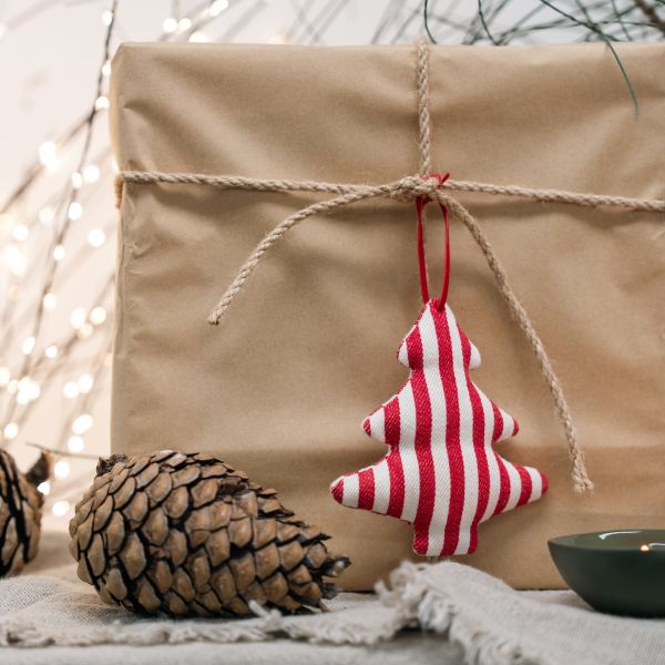 A red and white striped fabric Christmas tree ornament hangs from a rope tied around a brown kraft paper gift box, styled with pinecones and warm fairy lights in the background.