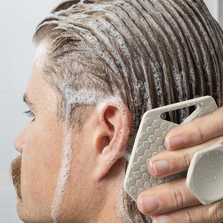 Person applying shampoo to their hair with a scrubber against a neutral background