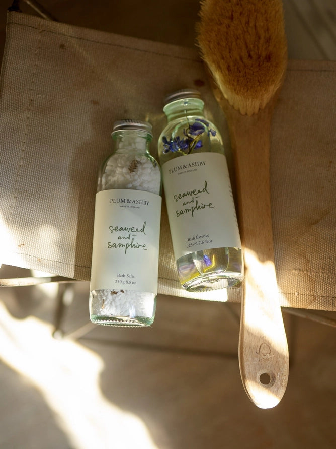 Two bottles of Plum & Ashby seaweed and samphire bath salts on a textured surface with a brush.